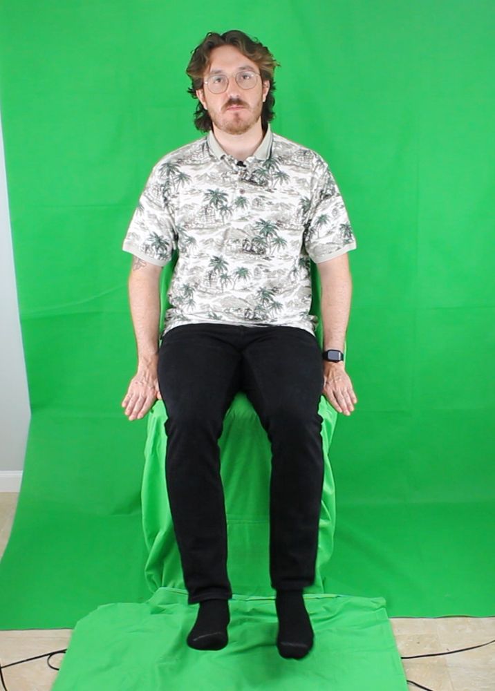tomatoanus sitting in a chair in front of a green screen. the chair is also covered in green to be keyed out later. feet dangling off floor.