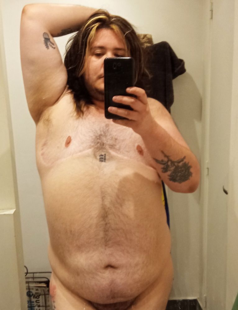 Nsfw - une photo de nu dans un miroir d'une personne non binaire transmasculine blanche, grosse, blanche. Il a les cheveux longs bruns avec des mèches blondes. Il a plusieurs tatouages, dont une chrysanthème sur le bras qui est visible sur la photo, et un lettrage sur le plexus solaire. Sur son torse il y a des cicatrices blanches de mammectomie. 