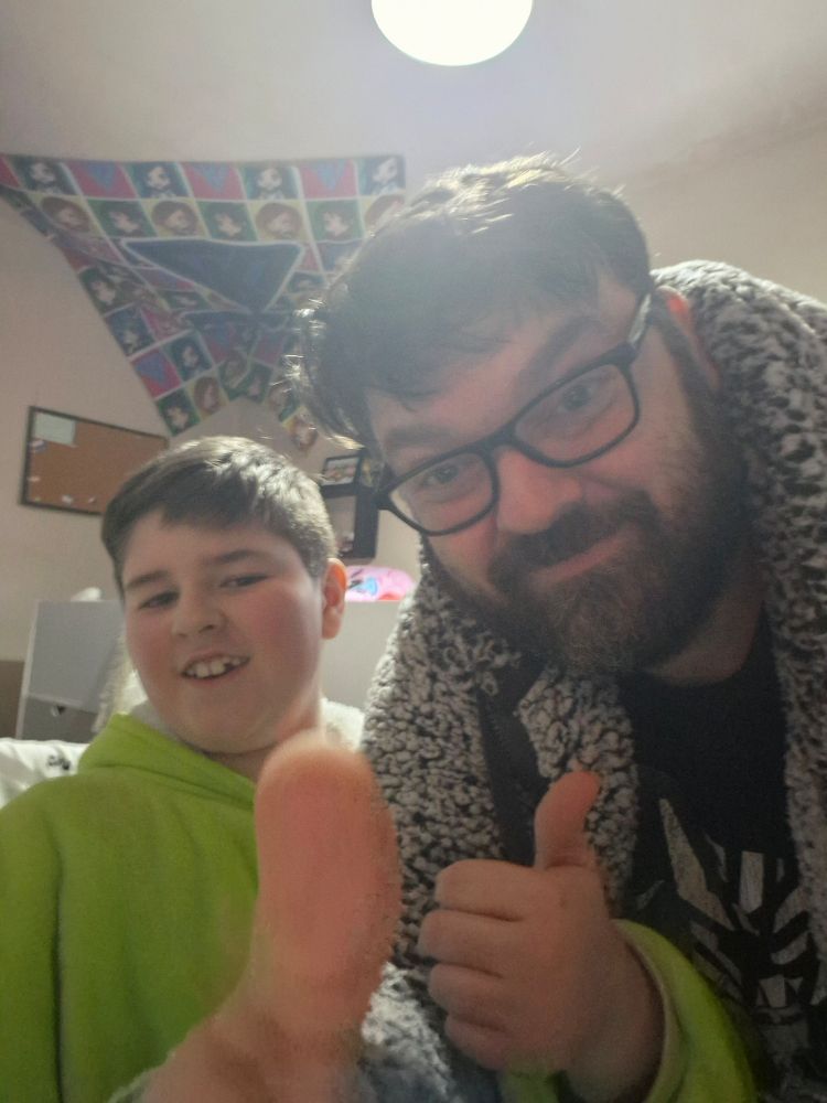 A boy and an oversized man child in warm looking clothes give a thumbs up each toward the camera. 