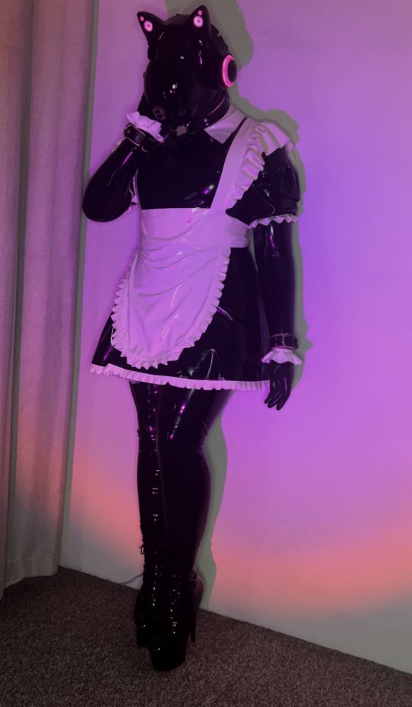 A latex clad entity stands up against a wall. It's latex maid dress implies it is set to do domestic duties. It has one hand up against its headset as it really enjoys the binaural beats that play while it does it chores. 