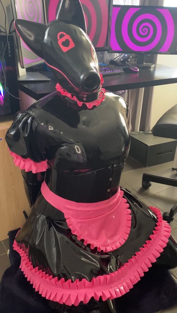 A latex coated entity kneels in front of a computer and desk. It is wearing a black and pink maid dress with its arms behind its back. The puppy hood it wears looks up to the user as if pleading for attention. Behind it are 3 monitors with black and pink spirals