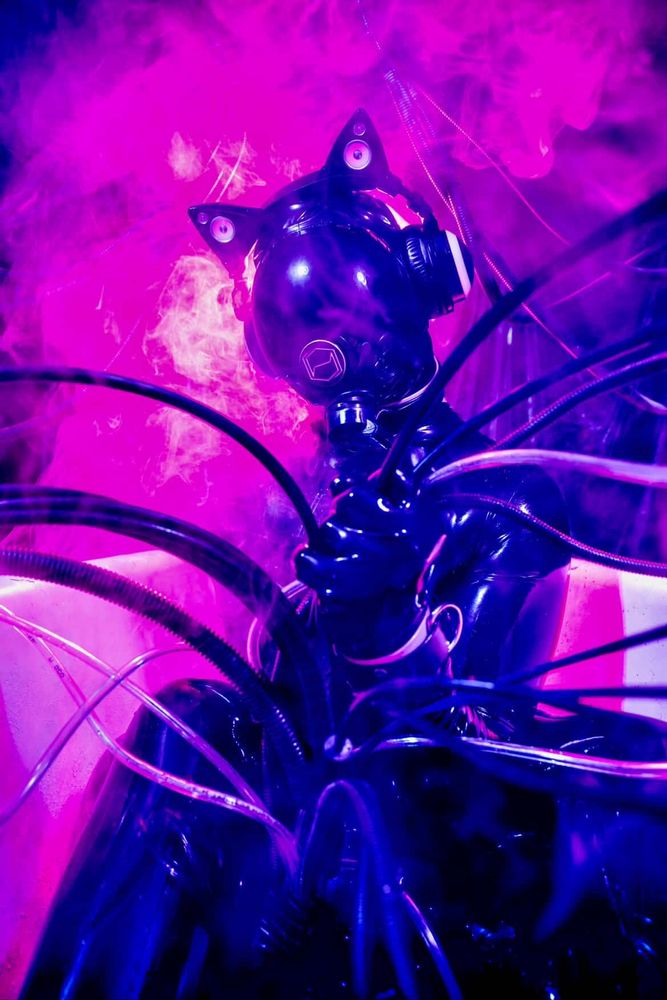 A Latex clad entity holding a mass of cables in tubes in its palm. Behind the gasmask and cat ear headphones it wears, pink smoke plumes behind it with wisps dancing across the drones visor.