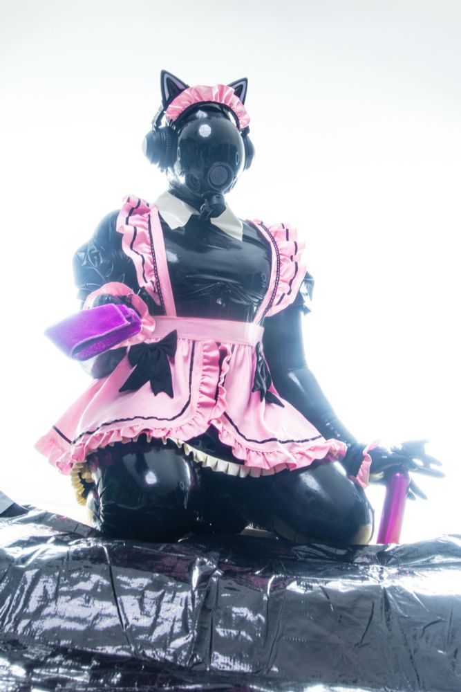 The entity nails down on her reflective surface holding a purple microfibre cloth in one hand and spray bottle. In the other. They are both purple. It is dressed in a rubbery made outfit with cat ear headphones and a gasmask for protection. 