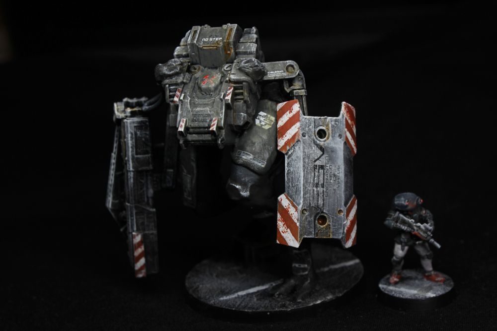 A painted model of a military robot, with a flat tank-like hull on very long, organic-looking legs.
It carries a shield and a weird weapon case in its crude arms.
The hull is dull olive green, the shield has white and orange stripes on its corners.
There is a humanoid figure next to it for scale, about 3 times shorter.