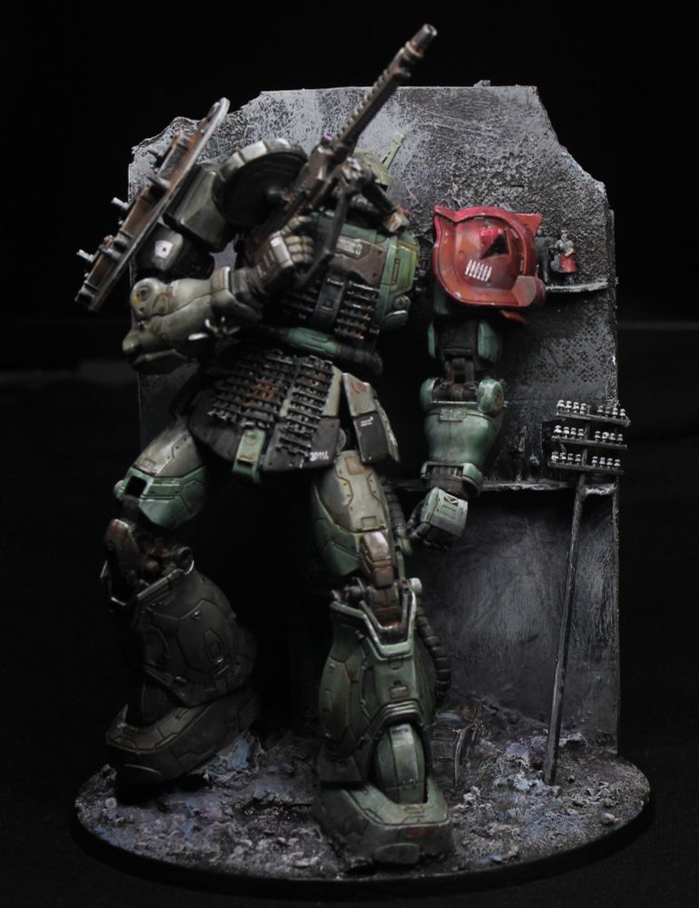 A large, pale green mech (Zaku) with a red spiky shoulderpad taking cover next to a large ruined wall.
There is a utility pole leaning onto the ruin, and some civilians standing right next to the mech.
The mech has slightly mismatched colours and improvised armour made of tank treads.