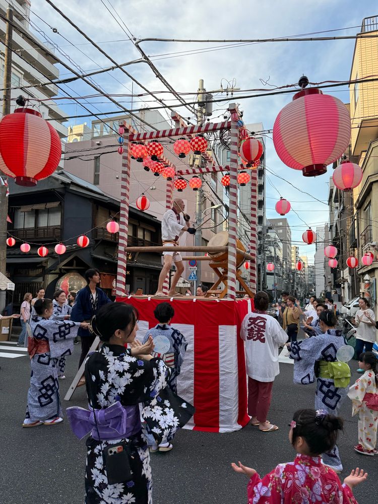Des femmes et des enfants (en kimono pour la plupart) dansent autour d’une petite estrade rouge et blanche sur laquelle est juchée en hauteur un papi et son taiko (tambour traditionnel). L’estrade est placée à un carrefour, en pleine rue, dans un quartier de Tokyo.