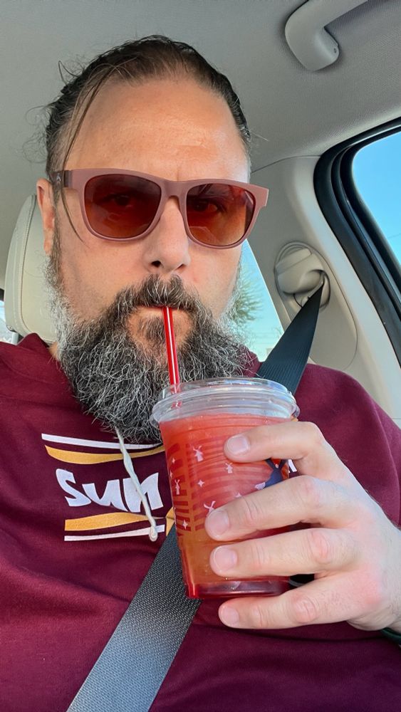 Tim Agne, a bearded man with his hair pulled back, wearing brown sunglasses and a retro Arizona State University hoodie, drinking a blended Pitchfork Rebel from Dutch Bros.