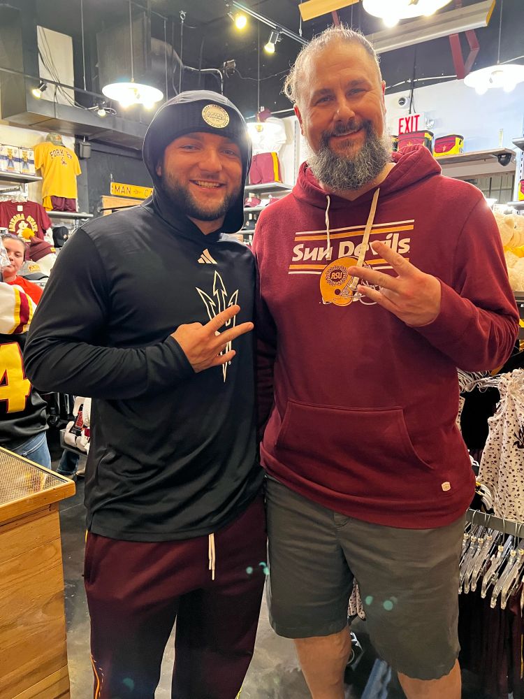 Arizona State University Sun Devil Football running back Cam Skattebo posing for a photo with Tim Agne in a store filled with ASU apparel. Both men are making the ASU pitchfork gesture with their hands.