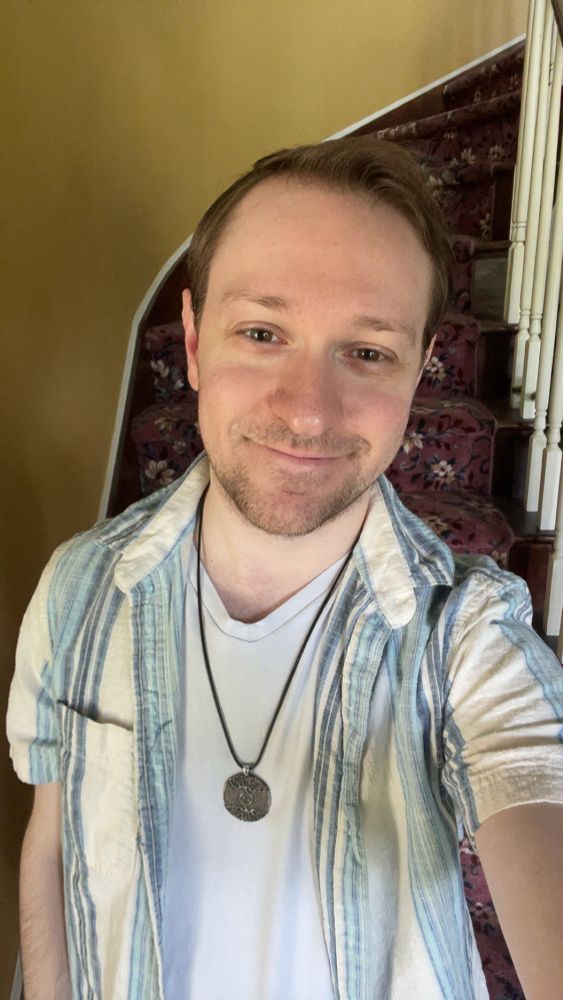 Me smiling and standing in front of a staircase with a pink floral pattern wearing a short sleeve with blue and white vertical lines button up unbuttoned over a white V-neck with a spiral tree necklace