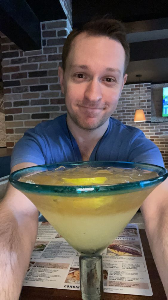 Me sitting at a restaurant table with my arms out and smiling while wearing a short sleeve V-neck shirt that’s blue at the top with a big margarita on the table between my arms 