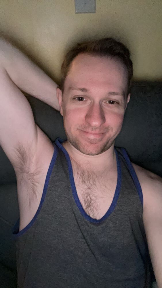 Me sitting on a couch and smiling while wearing a gray tank top with dark blue lining and with my left arm up behind my neck 