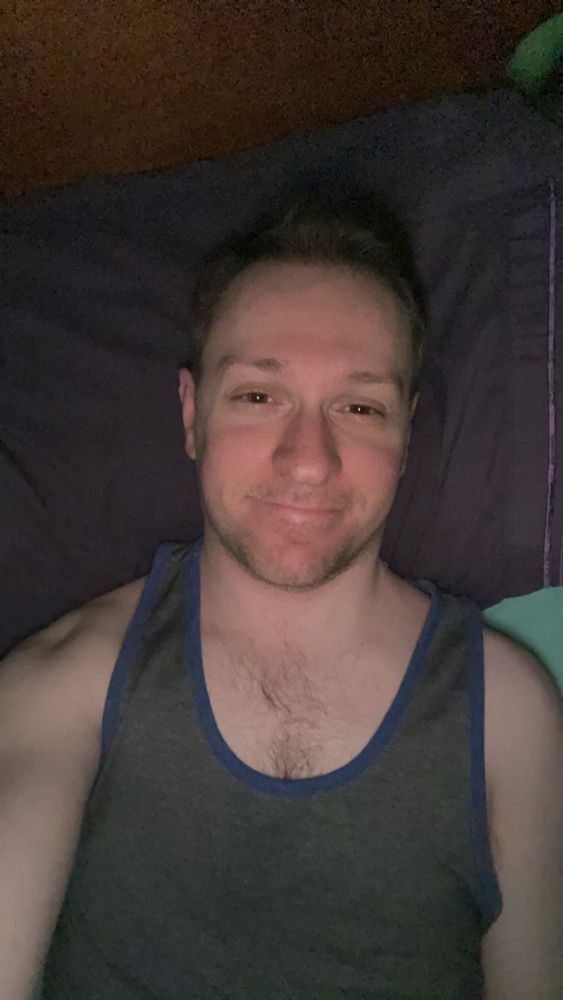 Me laying in bed and smiling on a pillow with a dark purple pillow case while wearing a gray tank top with dark blue lining 