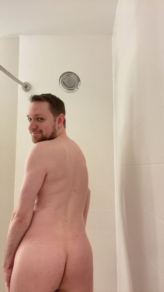 Me standing naked in a white tile shower facing away but with my head turned over my left shoulder with a smirk in the light