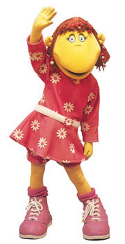Fizz from The Tweenies, a character with yellow skin, red hair and wearing a pink dress with white flowers and pink shoes with white laces. She is posing with one arm in the air.