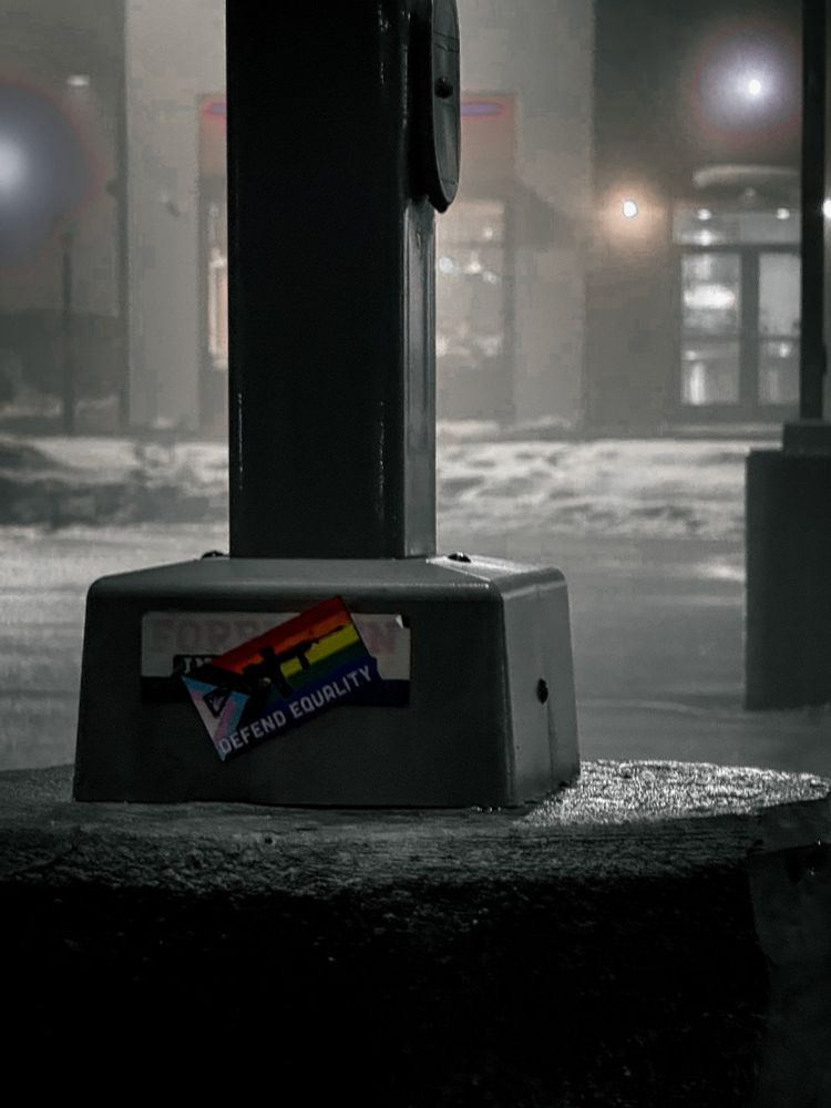 
A moody nighttime photograph featuring a metal pole on a city sidewalk. The base of the pole has a sticker with a rainbow flag, a silhouette of a weapon, and the words "DEFEND EQUALITY." The sticker partially covers another sticker beneath it. The background is blurred, showing a foggy street with illuminated windows and streetlights reflecting off wet pavement and snow piles. The image has a desaturated color scheme except for the rainbow flag on the sticker, which stands out.