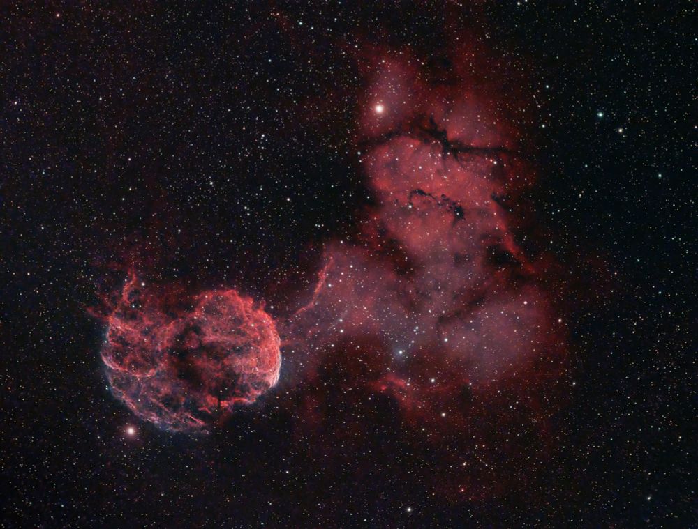 On the black background of outer space, a modeled pink nebulous cloud fills much of the center of the frame. Beginning about a quarter of the way from the left edge, a globular nebula with streamers of gaseous clouds is centered on the lower left "third" mark of the frame. To the right of that, expanding and extending upward to near the top of the frame and to the right to about a third of the way from the edge of frame, a more whispy, lighter nebulous cloud with darker pink edges and areas of very dark, making it look like a person with a headlamp on looking at the globular "jellyfish" nebula. The entire image is covered in white, yellow, red, and blue stars ranging in size from very small to medium. One larger medium star makes the "light" of the headlamp, and another larger medium star is just off the lower right of the jellyfish.