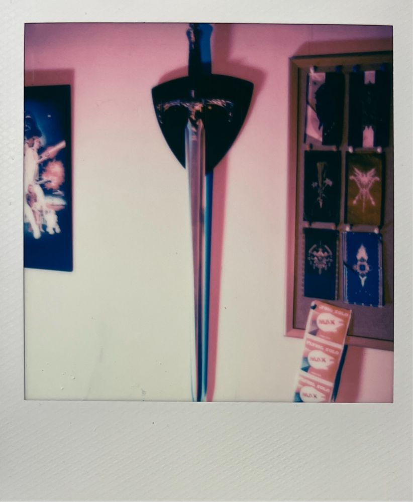 A sword hangs on a shield-shaped wooden wall mount, illuminated by blue and pink light. To the right, a cork board with banners. 