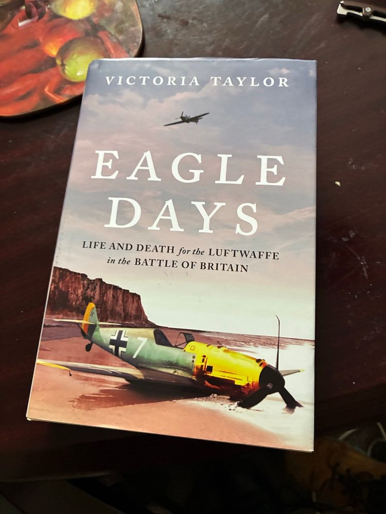 A picture of a book on a table: Eagle Days by Victoria Taylor: the cover shows a downed german single engine figther plane on what appears to be an english beach with step cliffs behind and a single engine figther plane in the sky above, presumeably a RAF figther plane.
Picture brazenly borrowed from Chris Sam.