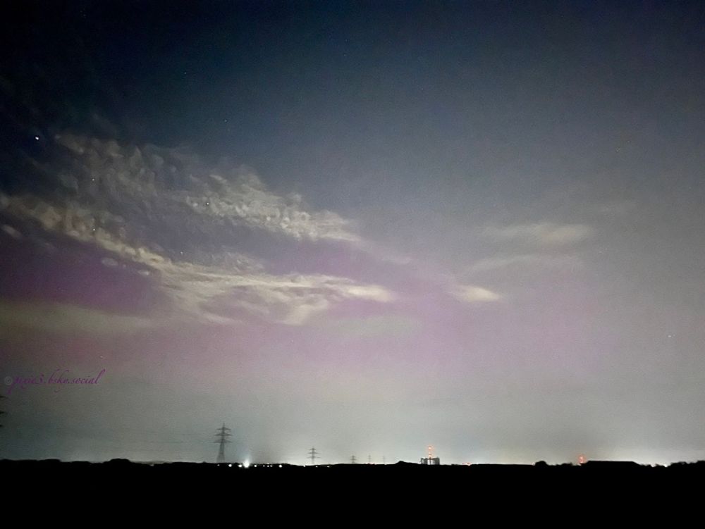 The night sky with some stars on the left side of the image, and some pink (in the middle of the picture) of an aurora borealis taking place that night. From the left side there are some fluffy traces of white clouds. The upper left corner is a darker blue.
On the ground there are dark fields and some silhouettes of electricity wires and towers plus some little light spots from a nearby village.
This aurora was nothing compared to the aurora from May 2024, but better than nothing, definitely!