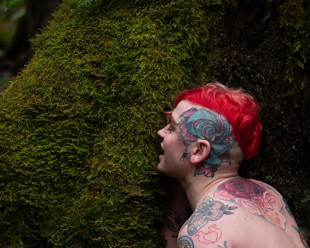 Person with tattoos and bright red hair leans into a patch of moss with an expression of joy and freedom 