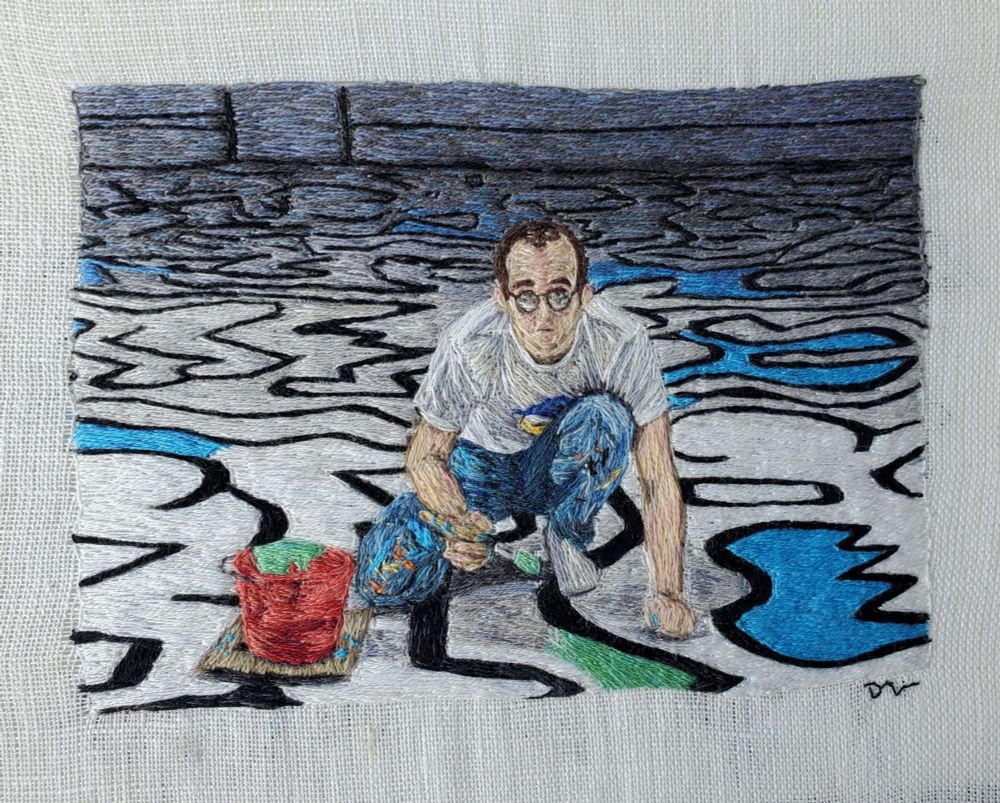 Embroidered thread painting of Keith Haring Painting Palladium backdrop. He's kneeling on the floor that is covered in white, black, and light blue squiggly lines. He is holding a paintbrush next to a red bucket and painting a small area green. It's stitched by hand on white linen fabric. 