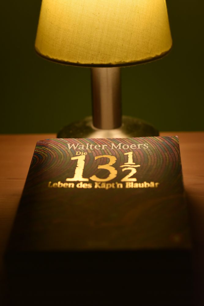 Im Vordergrund sieht man ein Buch auf einem Nachttisch aus Holz, welches von einer dahinter stehenden Lampe beleuchtet wird. Durch das Licht leuchtet der Titel "Die 13 1/2 Leben des Käpt´n Blaubär" golden auf.