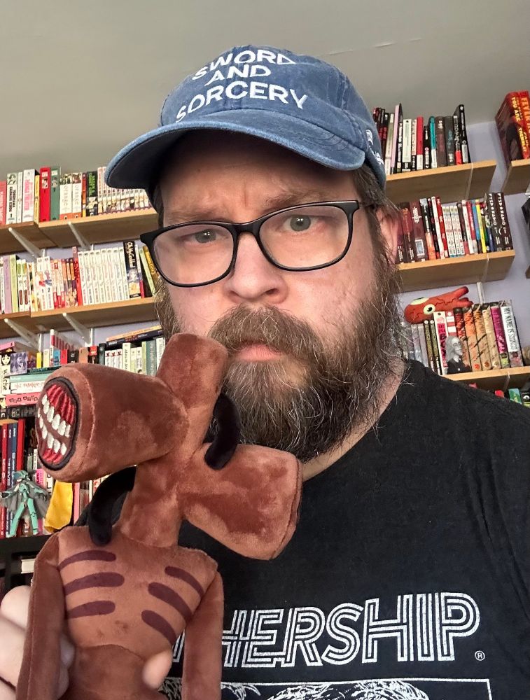 me, a guy with a beard wearing a black shirt and a baseball cap stands in front of some shelves of books holding a plush stuffed toy version of my character siren head