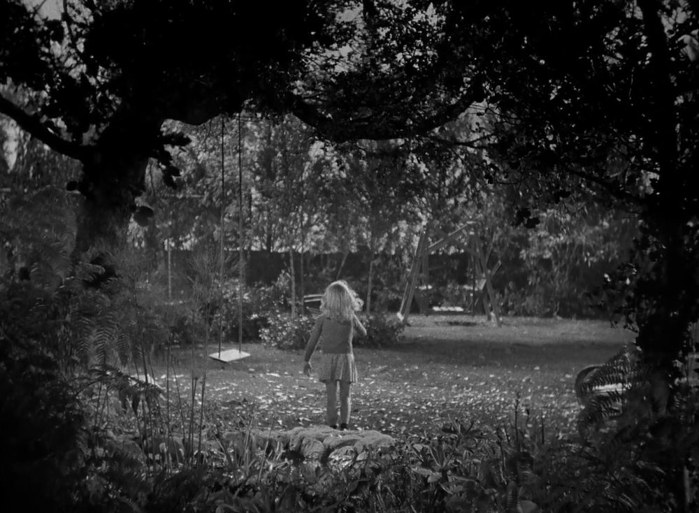 Amy, the lonely child protagonist of Curse of the Cat People stands in a lush backyard, framed on either side by trees