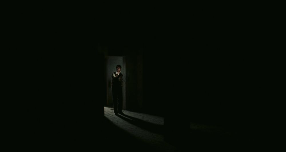 A man stands in the doorway of a cavernous, pitch-black room, his face lit by a single match, his shadow thrown out ahead of him.