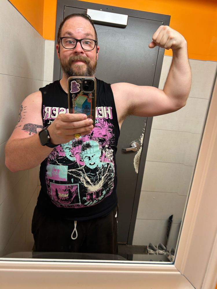 It’s Trevor, a bearded white dude with glasses and a big forehead. He’s wearing a neon teal and pink hellraiser tank top and flexing in a gym bathroom. He has a tattoo of the mutant dog from the thing on his left arm