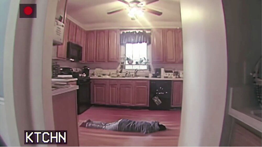 a screenshot from the short film THIS HOUSE HAS PEOPLE IN IT. a suburban kitchen, panelled in wood, as seen from a spy camera. the letters KTCHN are in the bottom left of the frame, and the angle is warped in the manner of a fish-eye lens. a woman in grey sweat shirt and sweat pants is slowly sinking into the floor in an unnatural and uncanny way