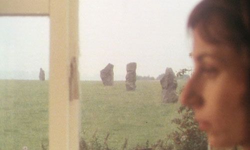 a woman sits in profile in the foreground. in the background, a field with some large standing stones. An image from STIGMA (1977)
