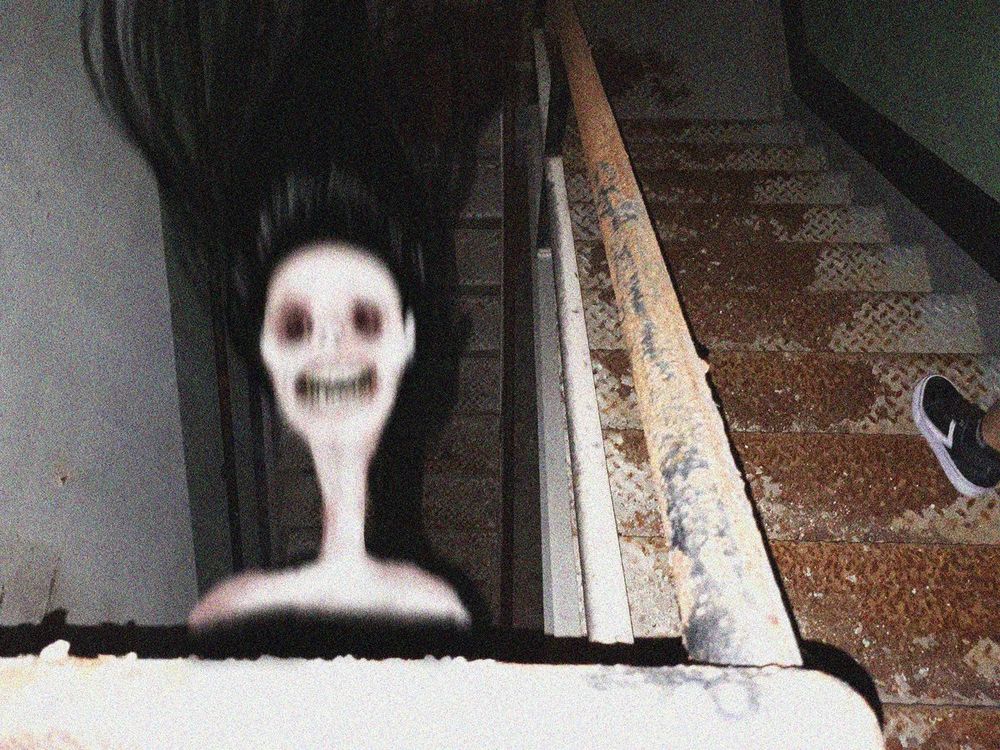 a photo taken looking down a stairwell in a dirty and abandoned building. to the right, on the stairs, a single shoe and part of a shin are visible. jutting forward into view over the downward stairway is a pale woman's face with an abnormally long neck, her black hair spread out. she appears to have been caught in motion, her mouth open in a cry. both of her eyes have been gouged out.
