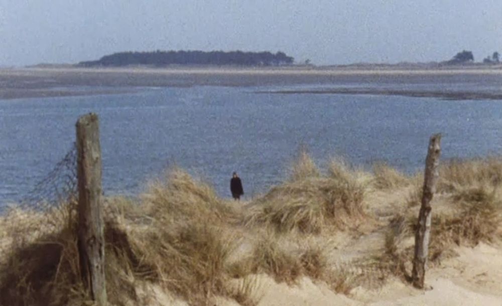 a body of water, with a slope of sand and brush leading towards the viewer. an ominous figure in black stands by the water. he's staring at us. a still from A WARNING TO THE CURIOUS (1972)