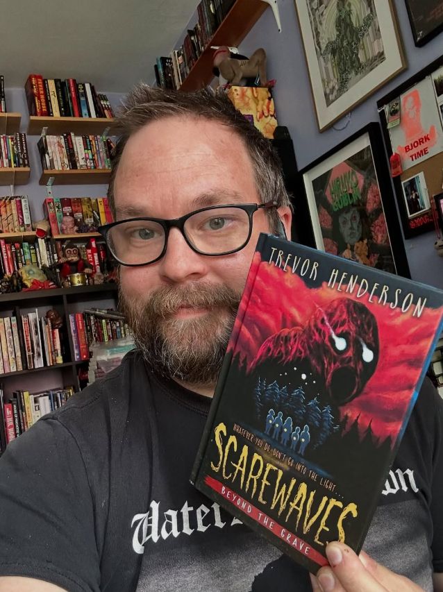 it's me, a guy with a beard and glasses, holding a copy of scarewaves: beyond the grave. there's a shelf full of books behind him