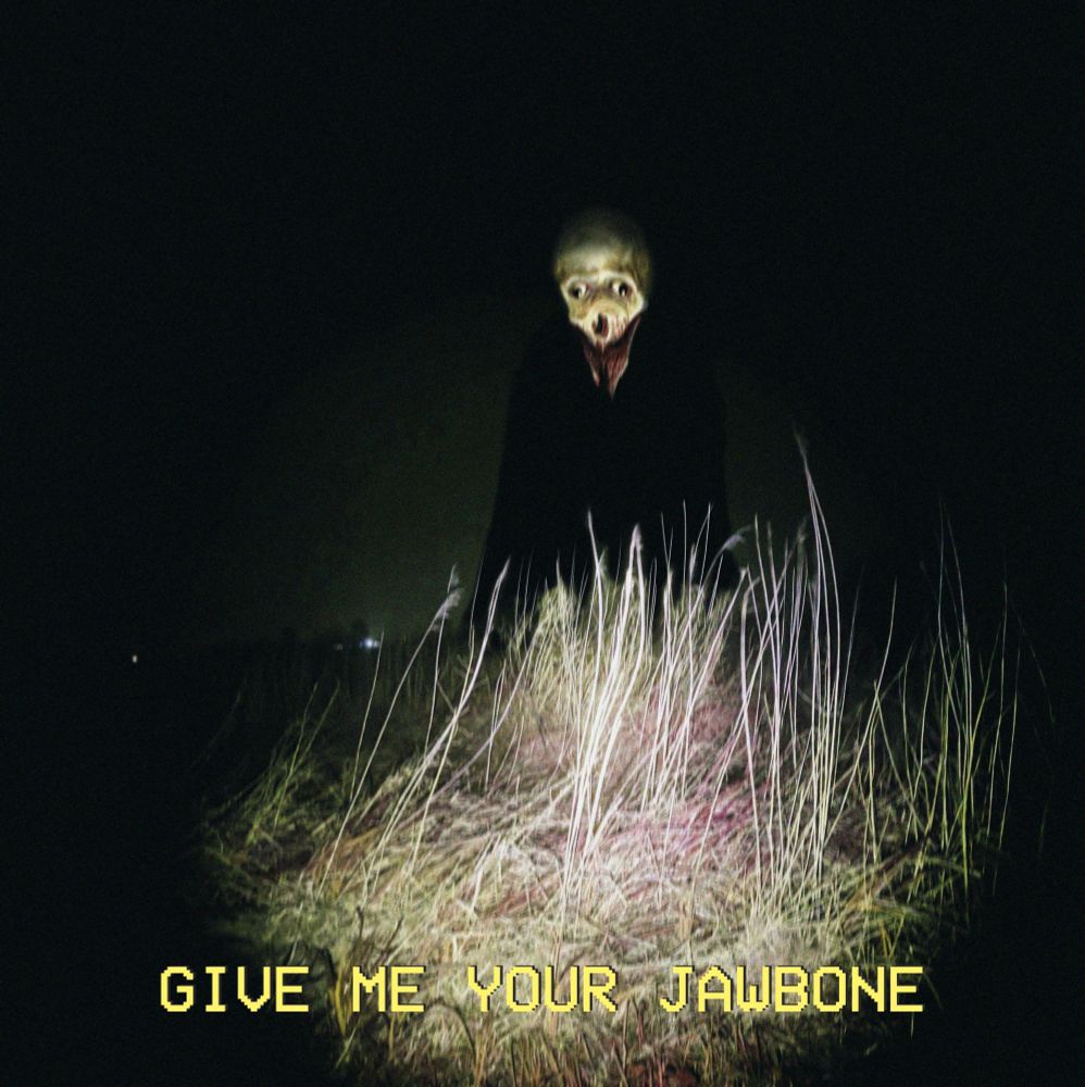 a skeletal figure standing amongst the tall grass at night, in the light of a flashlight. he's missing his lower jaw. yellow text on the bottom of the image says "give me your jawbone"