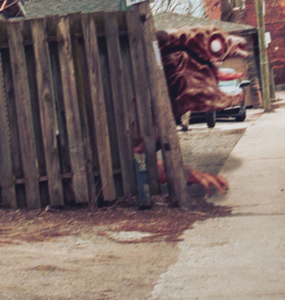 A city alley with a broken down wooden fence. An animal like a flayed calf walks into view from behind the fence