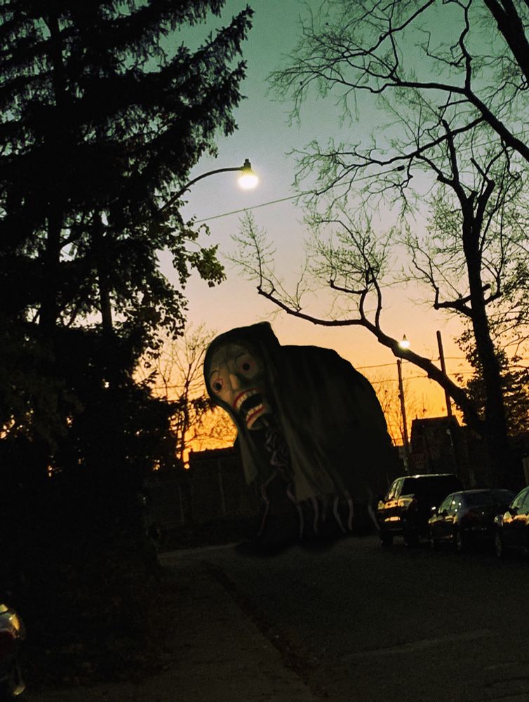 A suburban street at sunset. Something or a bunch of somethings under a giant cloak hoist a giant wooden head, as part of some ritual or parade.