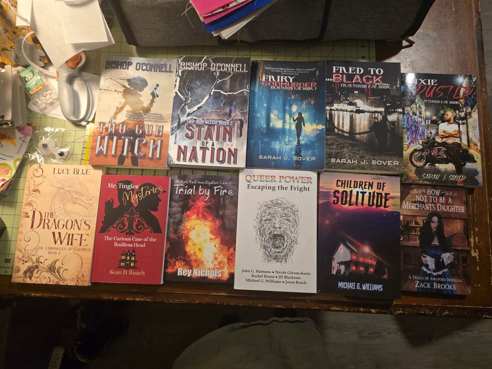 Selection of 11 books on a table. Titles include Two-Gun Witch, Stain of a Nation, Fairy Godmurder, Faed to Black, Pixie Dusted, The Dragon's Wife, Mr. Tingles Mysteries, Trial by Fire, Queen Power, Children of Solitude, and How Not to Be a Merchant's Daughter. 