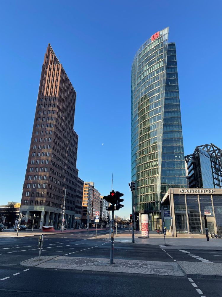 Zwei Hochhäuser am Potsdamer Platz in Berlin. Eine Fußgängerampel zeigt auf Rot (Warten). Rechts zeigt sich ein Vorbau aus Glas des Bahnhofes, an dem oben der Schriftzug BAHNHOF zu sehen ist. 