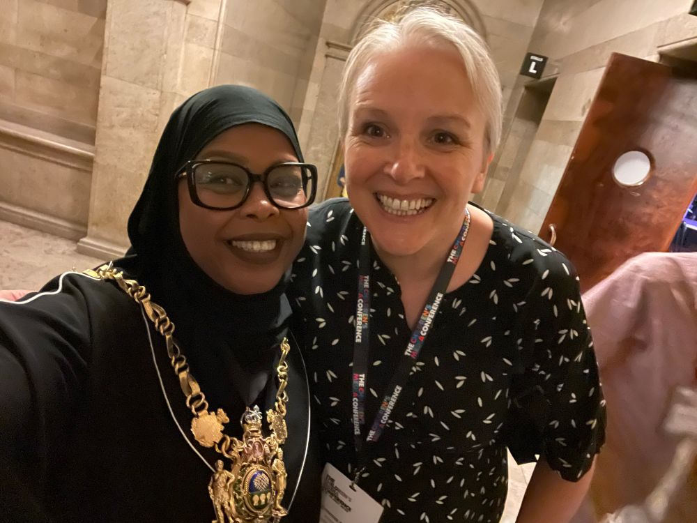 A photo of two women one is black, wearing a black headdress and wearing a large gold ceremonial necklace, the other is white with short grey hair, both are smiling a lot 