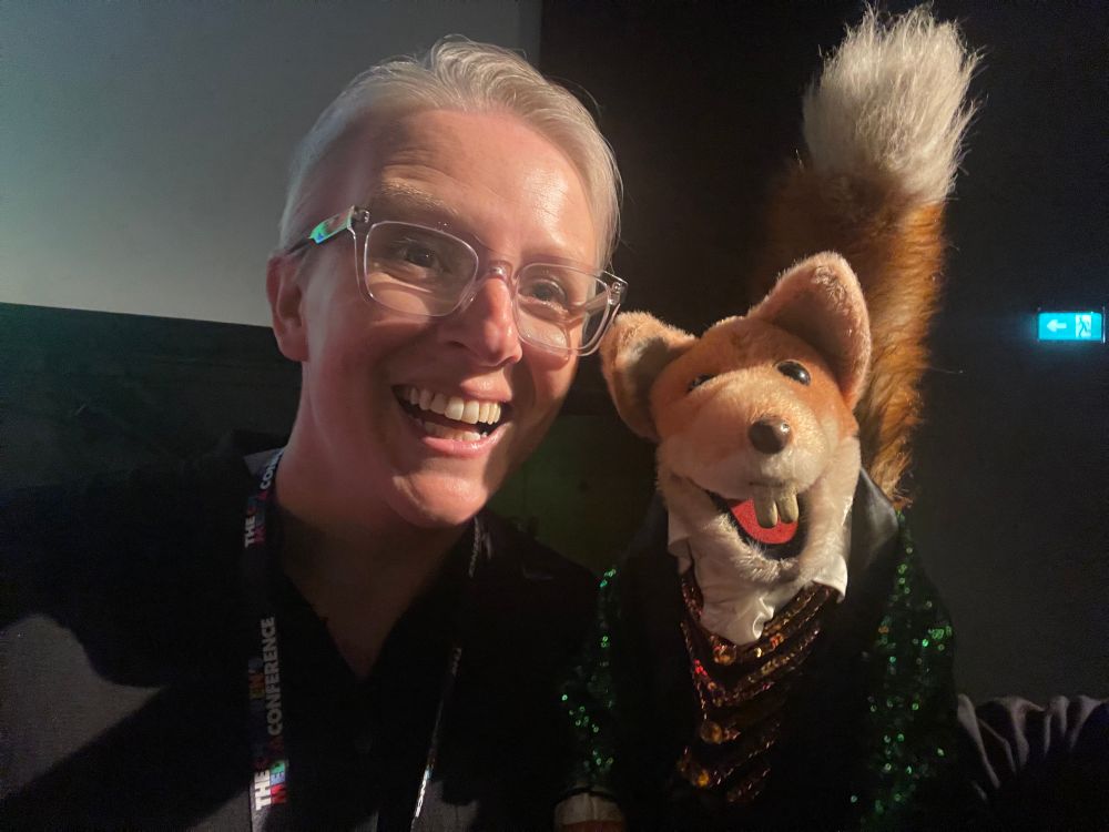 A photo of a middle aged white woman with short grey hair and glasses grinning like she was 7 years old next to the children’s media mega star Basil Brush who is a a red fox wearing a green sequin jacket