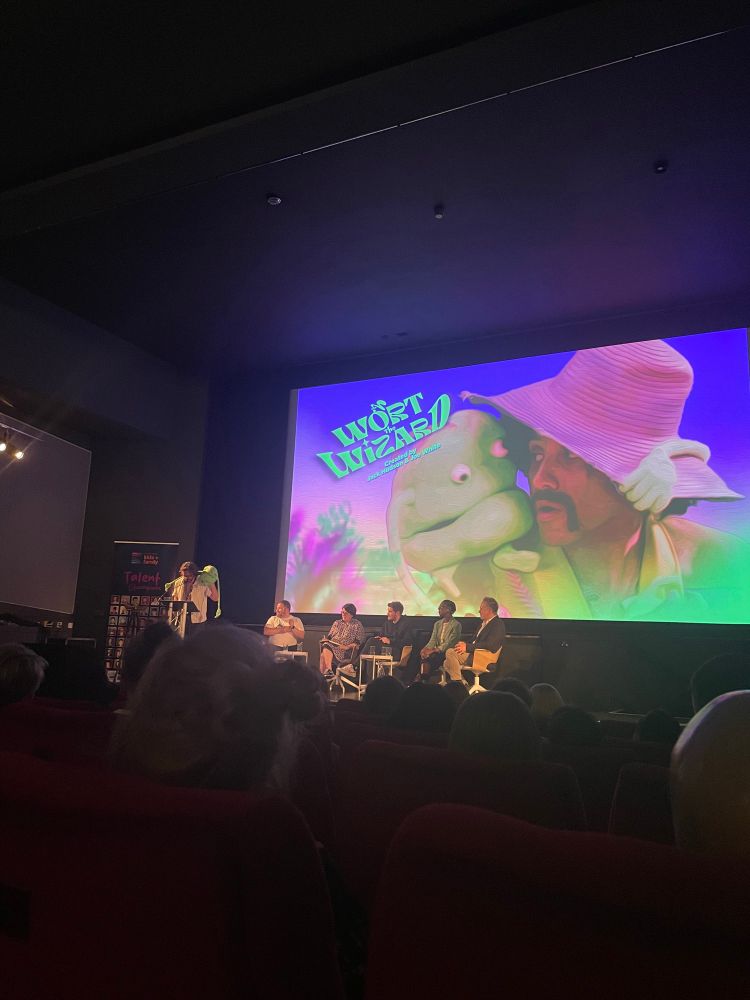 A picture of a man on stage with a green blob puppet on the screen behind him the is an image of a man in a straw hat with a questionable moustache- the puppet is on his shoulder the text reads Wart The Wizard Created by Jack Hudson & Joe White