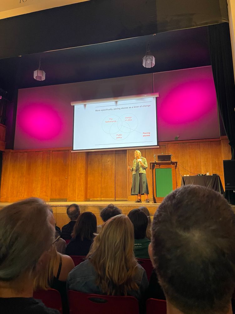 A woman on stage her slide has a Venn diagram with “Netherlands” “Design of Cities” and “Public policy” on the outside the centre is labelled “Paving stones”