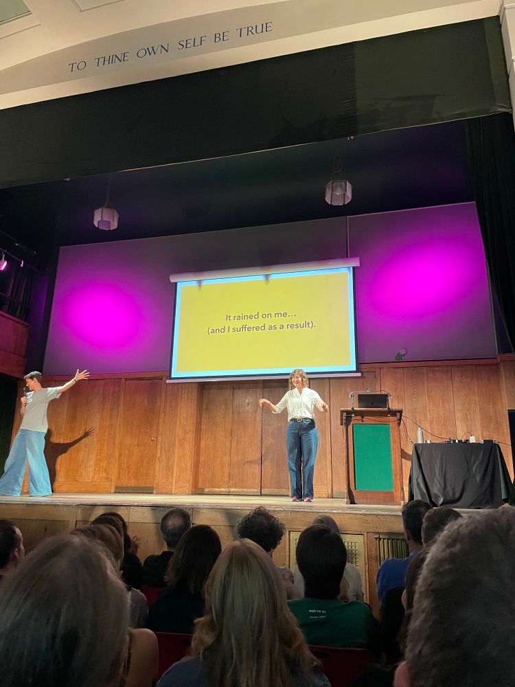 A woman speaking on stage, a slide behind her reads “It rained on me … (and I suffered as a result)