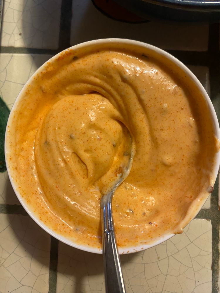 Bang bang sauce, orange in color with little chili flakes in a white dish and a silver spoon.