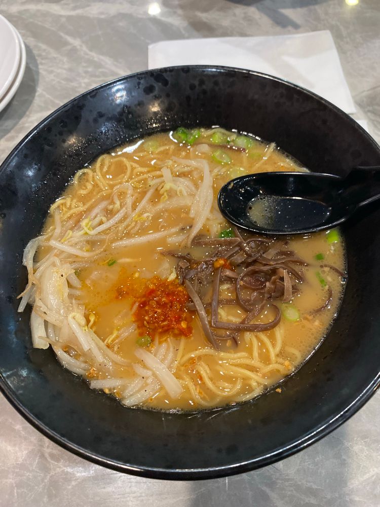 Black bowl filled with golden broth, ramen noodles, chili crisp hot sauce and green onion. A black spoon is resting on the side of the bowl.
