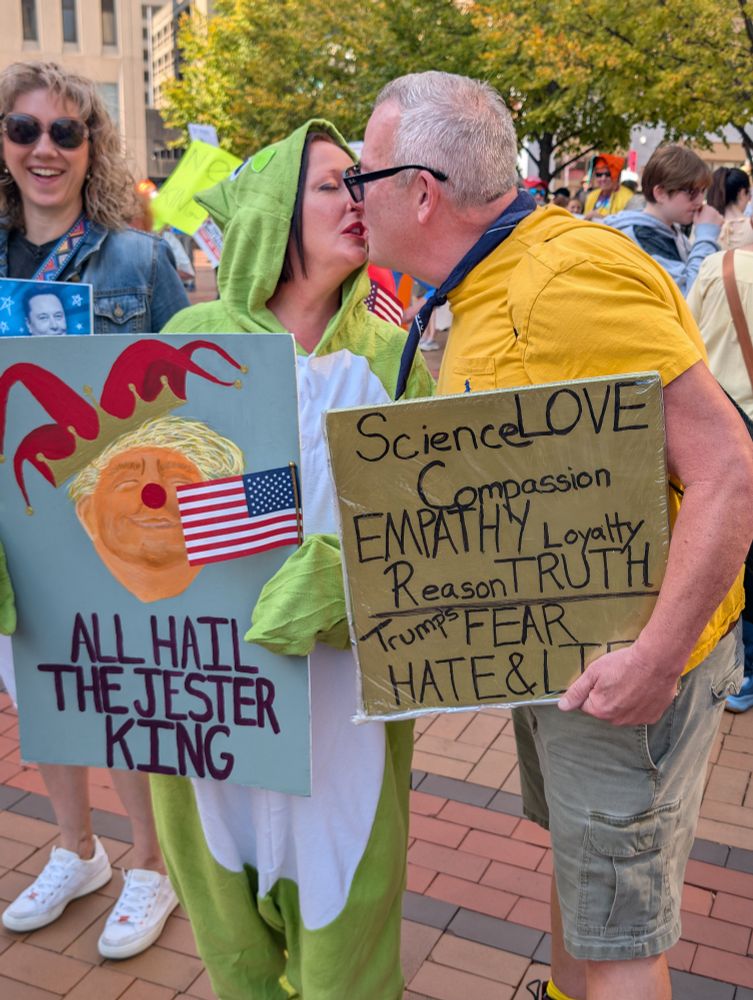 Woman in a frog suit kisses a man in a yellow Tee. 
She holds a sign that says: All hail the jester king and his sign says science love compassion empathy reason truth trumps fear hate & lies. 