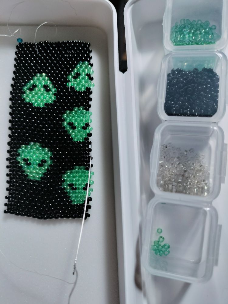 Even count peyote stitched alien faces. The background is black beads, the aliens are green.