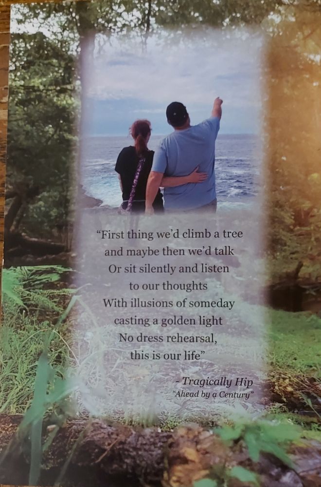 The back of the memorial card for my honey. The background is wilderness, and the  foreground is a picture of him and me. We are facing away from the camera, looking out to sea. It is a picture from our trip out to Halifax in 2024. Beneath the photo, the lyrics of the first verse of the Tragically Hip's song "Ahead by a Century" are printed.